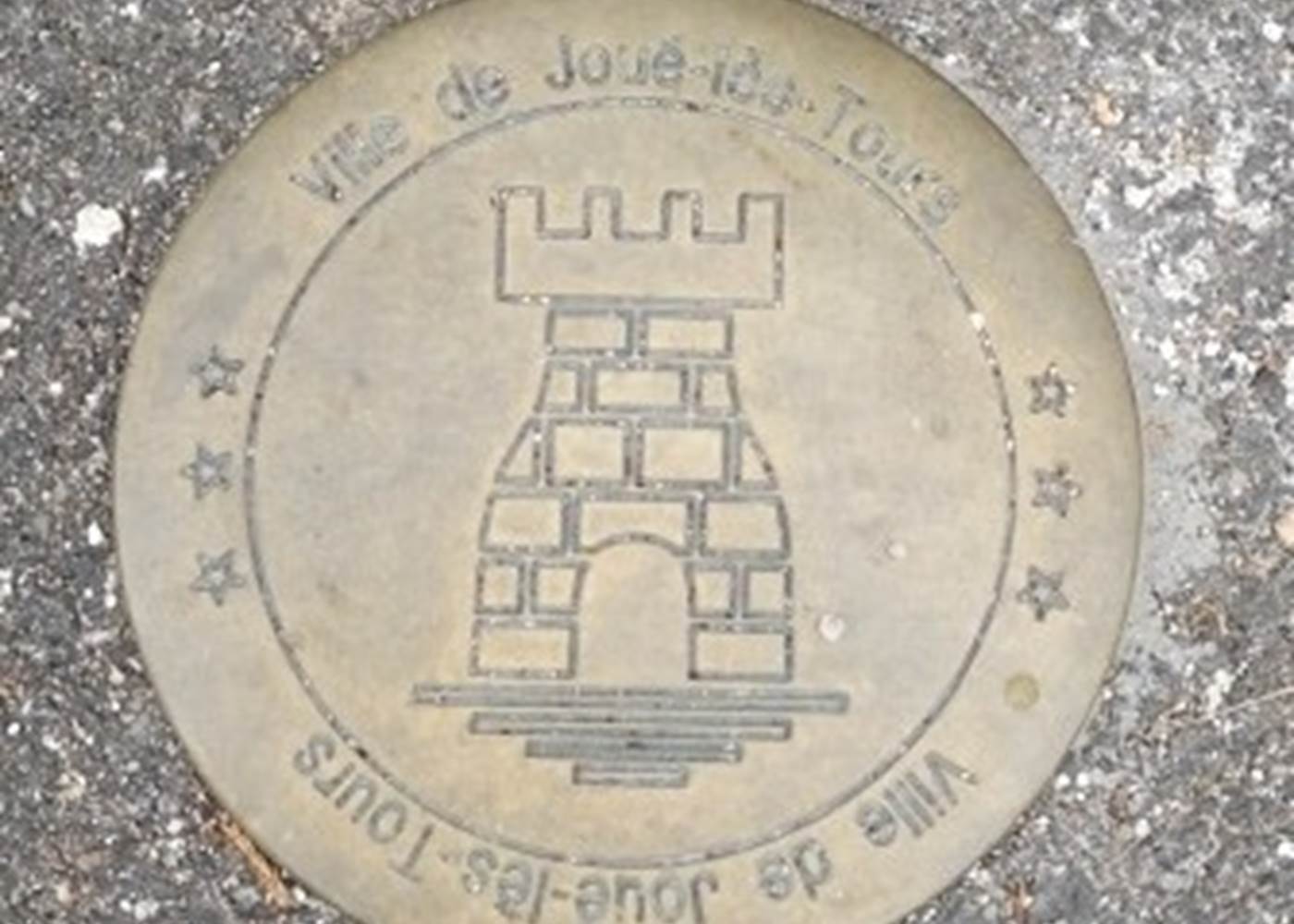 Ecusson des Monuments Historiques de la Ville de Joué-Lès-Tours incrusté dans le trottoir devant le portail piétons