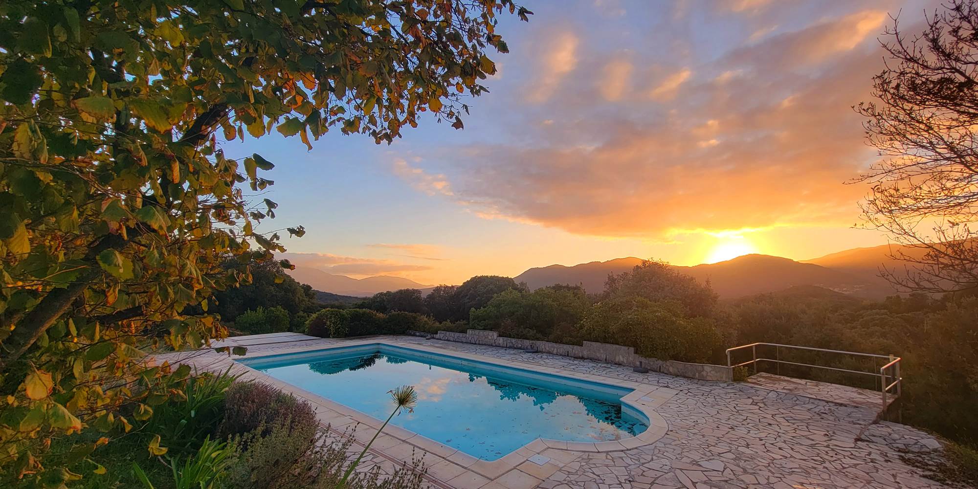 Piscine avec vue panoramique sur les montagnes corses embrasées par le soleil couchant.