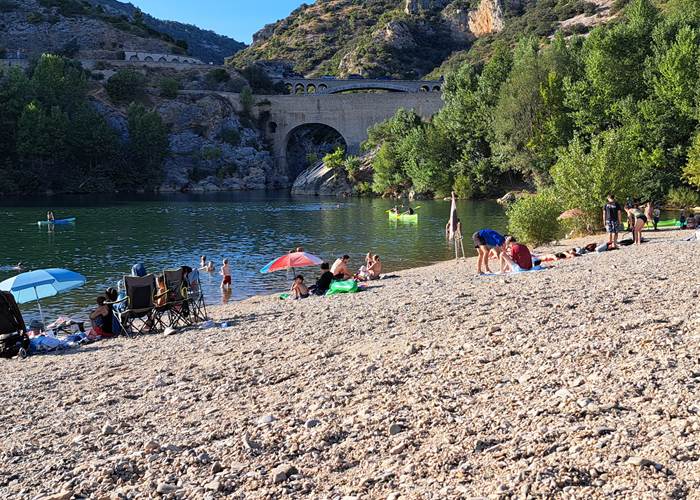 Plage de baignade sur les rives de l'Hérault, avec des familles, des canoës et le pont du Diable au fond des gorges.